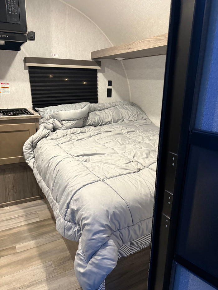 RV rental interior: A compact bedroom with a grey bedspread, small kitchen counter, stove, microwave, and wood flooring.