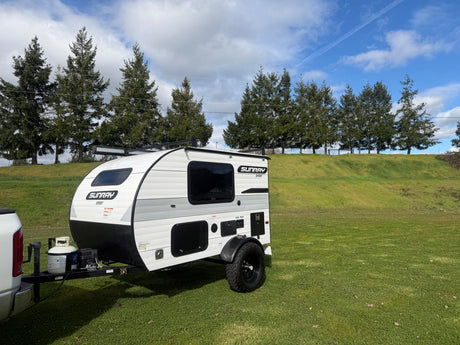 This Sunray Sport teardrop rv rental, hitched to a truck, awaits adventure on a lush green field under a blue sky.
