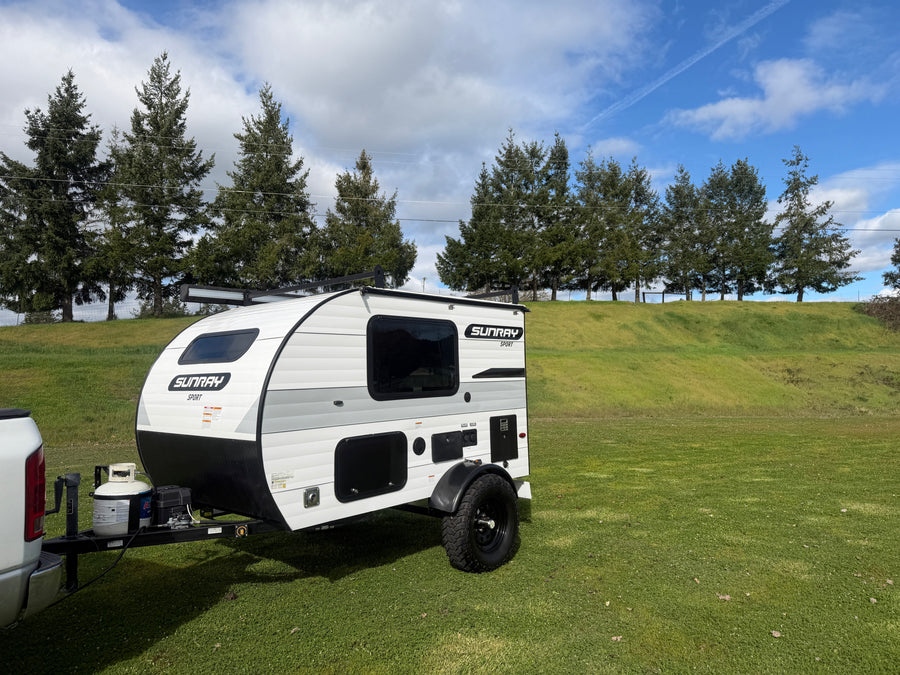 This Sunray Sport teardrop rv rental, hitched to a truck, awaits adventure on a lush green field under a blue sky.