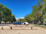 Blue SUV with a teardrop RV rental at a scenic campsite, flanked by trees with snow-capped mountains under a clear sky.