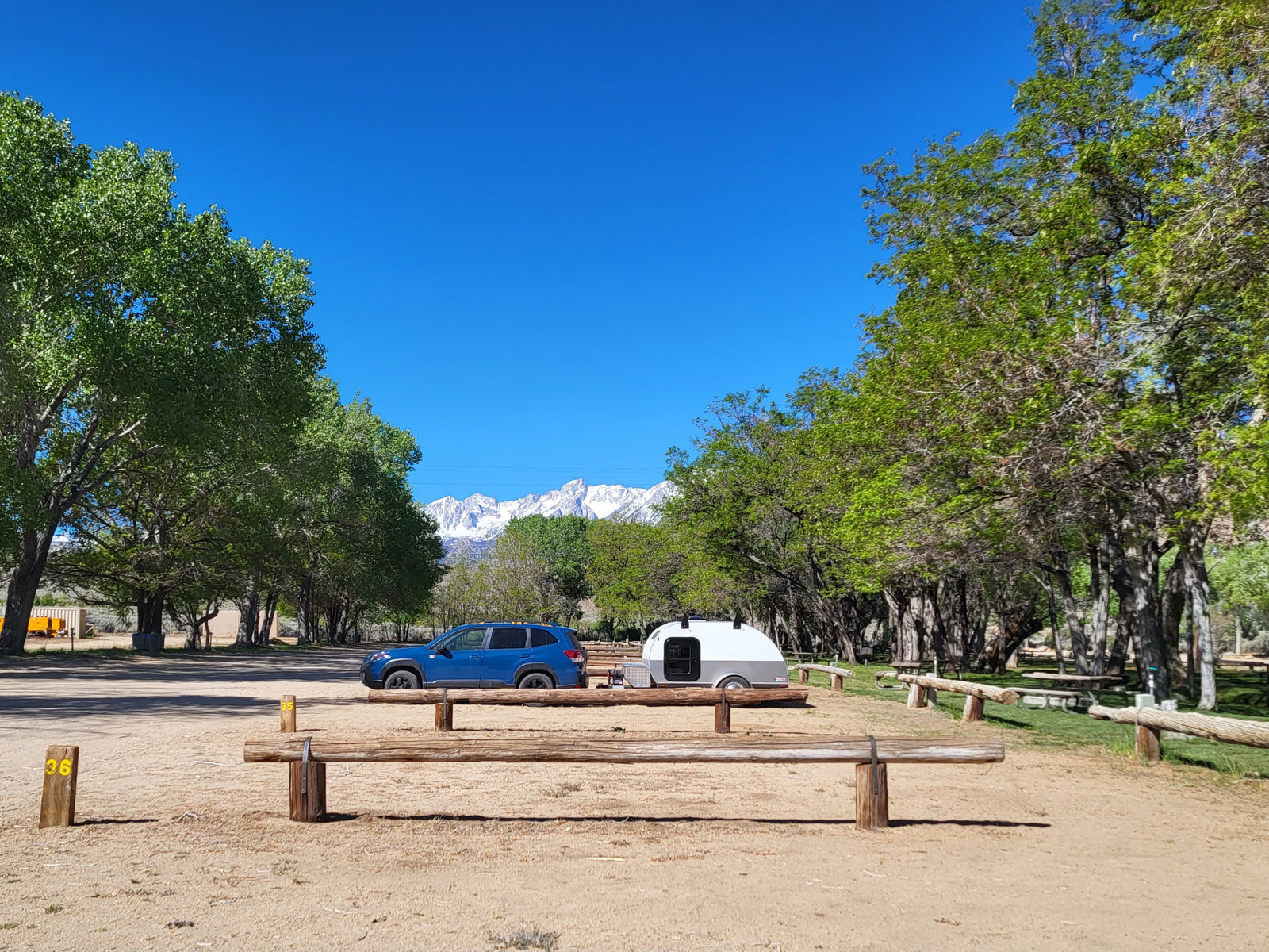 Blue SUV with a teardrop RV rental at a scenic campsite, flanked by trees with snow-capped mountains under a clear sky.