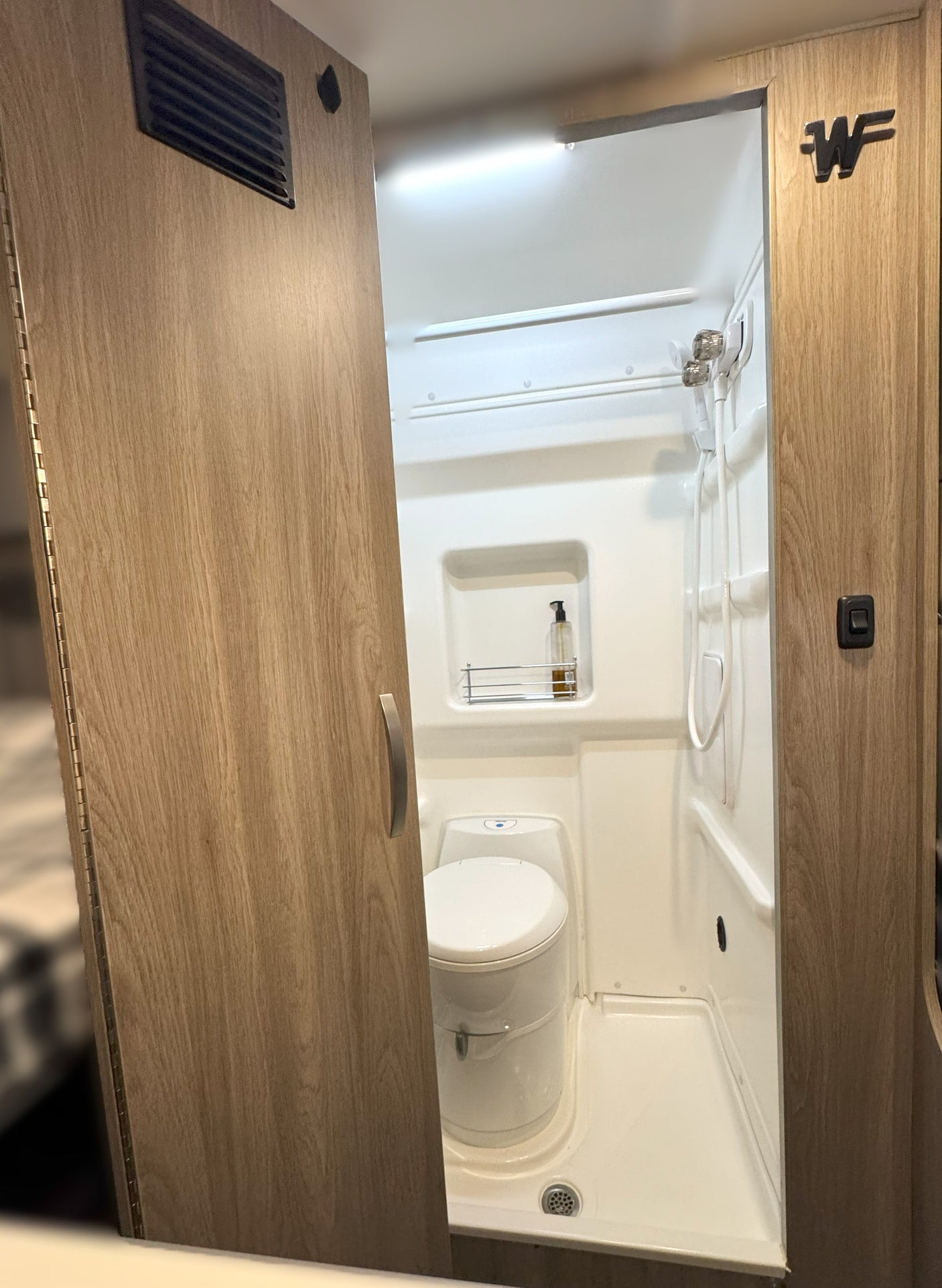 Explore this RV rental's compact wet bath, featuring a toilet, shower, and soap shelf. A light-wood door stands ajar.