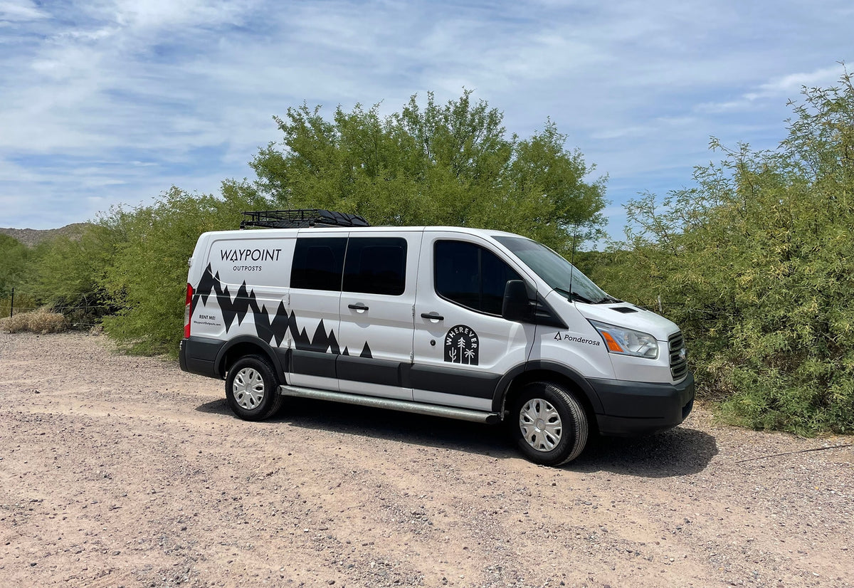 White Waypoint Outposts RV rental van, a Ford Transit with mountain graphics, parked on gravel amidst green desert plants under sunny skies.