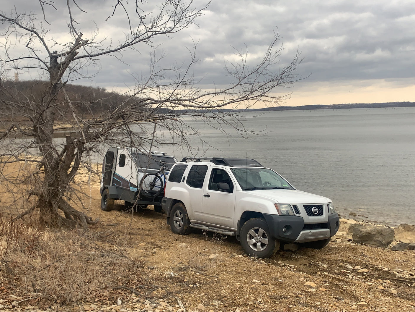 White SUV towing a compact RV rental camper with a bicycle, parked by a lake on a cloudy day. Ready for adventure.