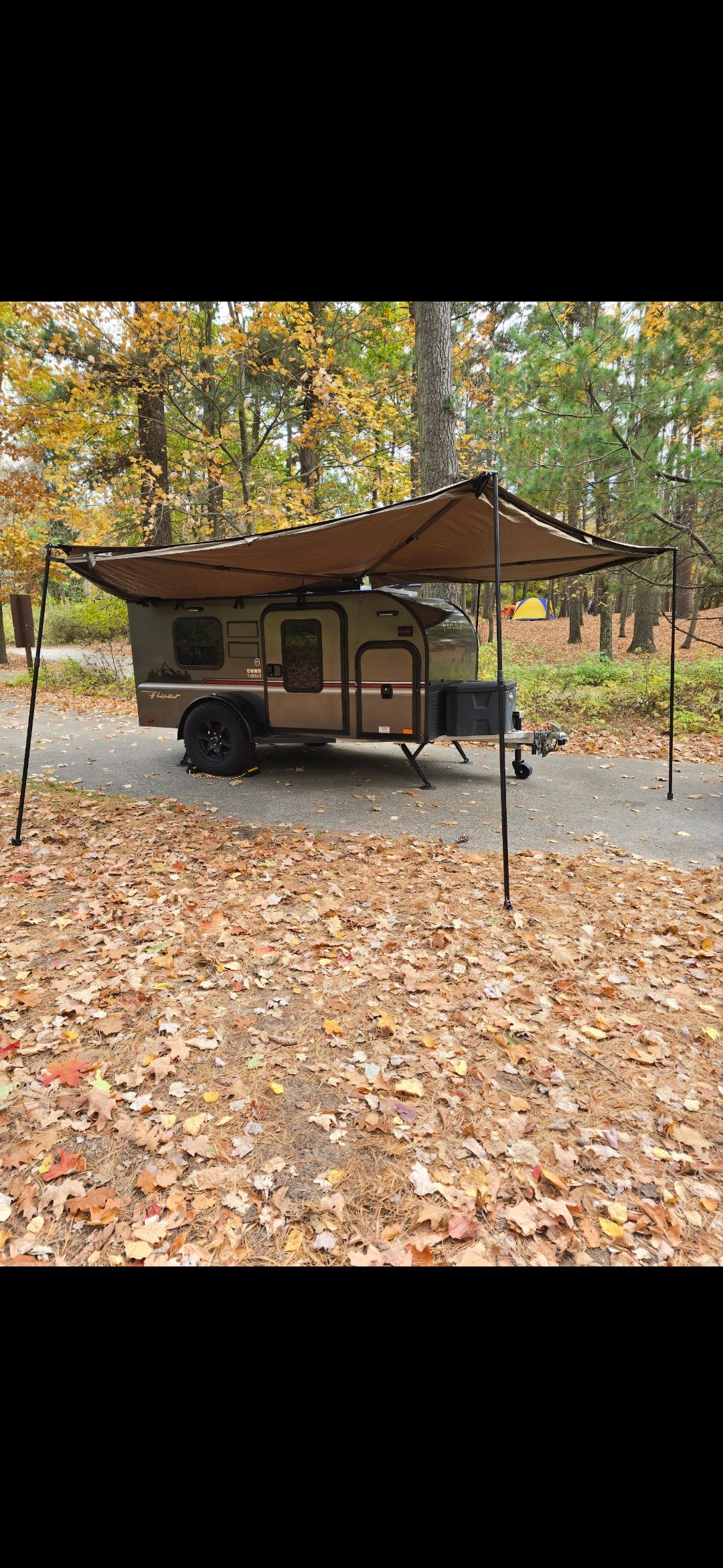 RV rental teardrop camper with awning, parked in a vibrant autumn forest campsite covered in fall leaves.