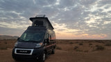 Dark gray Ram ProMaster RV rental parked in an expansive desert landscape under a cloudy sunset sky, pop-top extended.