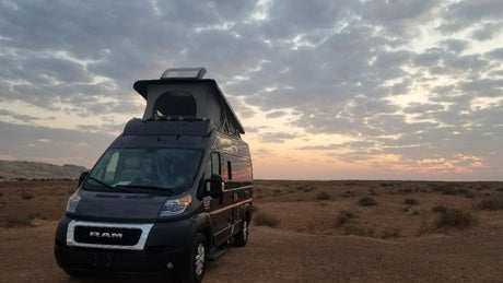 Dark gray Ram ProMaster RV rental parked in an expansive desert landscape under a cloudy sunset sky, pop-top extended.