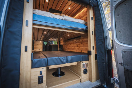 This RV rental van's wood-paneled interior shows a bunk bed & convertible lower bed. Ideal for mobile living.