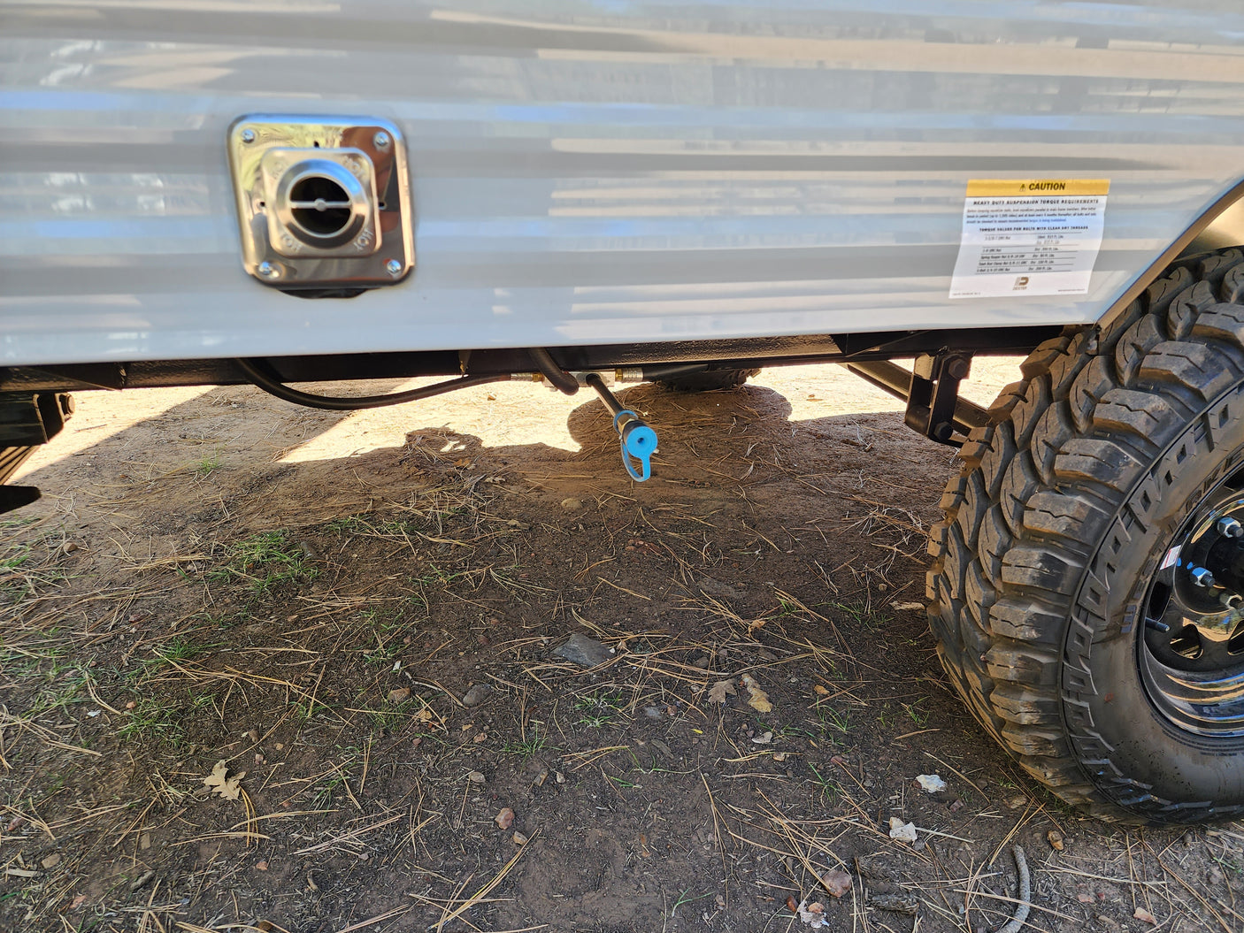 Side view of an RV rental: close-up of water connection, gas line, and rugged tire on dirt. Suspension caution label visible.