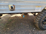 Side view of an RV rental: close-up of water connection, gas line, and rugged tire on dirt. Suspension caution label visible.