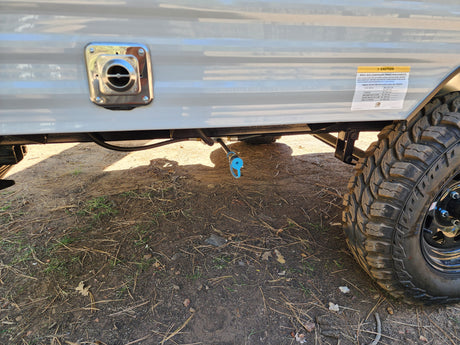 Side view of an RV rental: close-up of water connection, gas line, and rugged tire on dirt. Suspension caution label visible.
