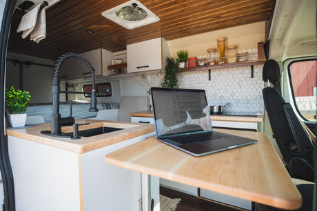 Sleek RV rental interior: kitchen with hexagonal tiles & black sink, laptop on a wood table, bed visible in back.