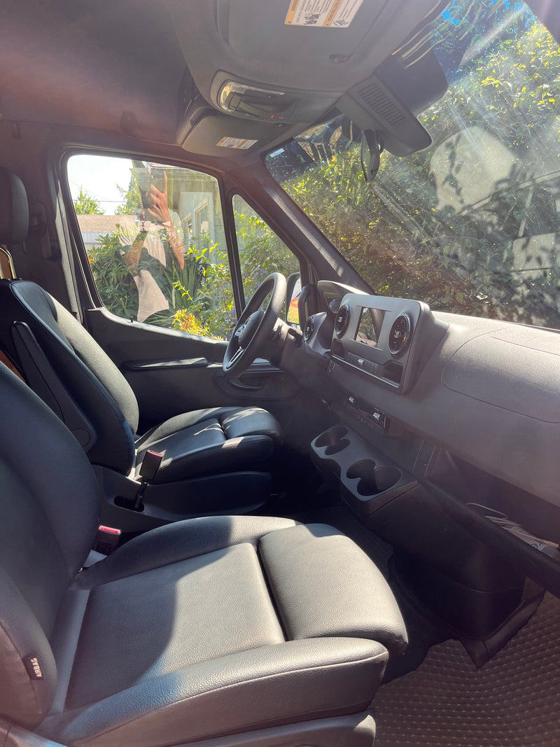 RV rental interior: black seats, dashboard, and steering wheel visible in the driver's area. Sunlit trees outside windshield. — photo 8