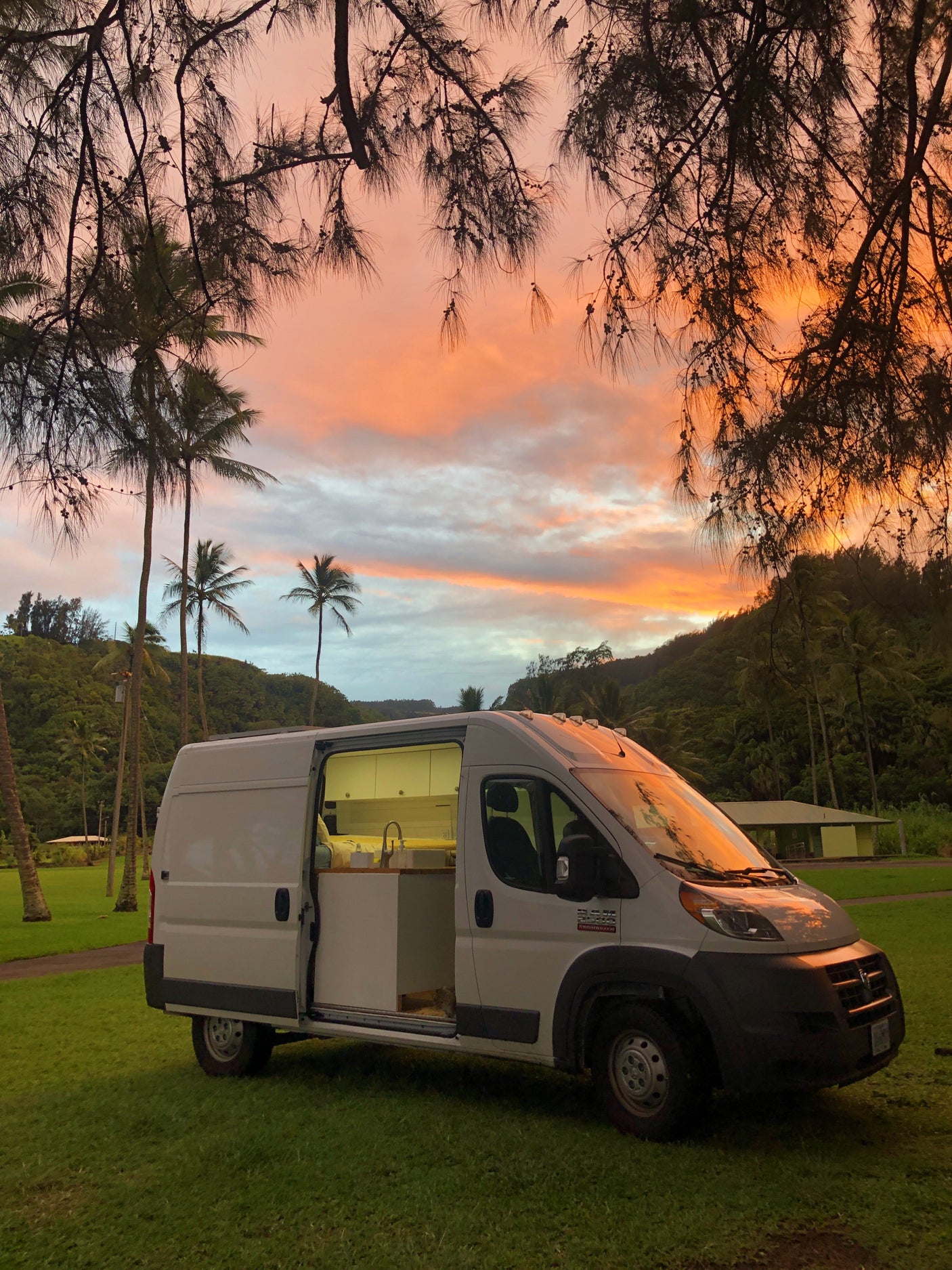 White rv rental camper van on grass, side door open to reveal interior. A vibrant orange and pink sunset illuminates tropical trees.