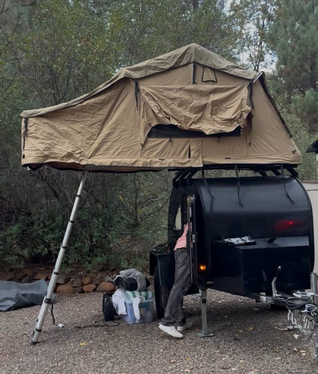 A unique rv rental experience: a rooftop tent deployed over a black teardrop camper trailer at a campsite.