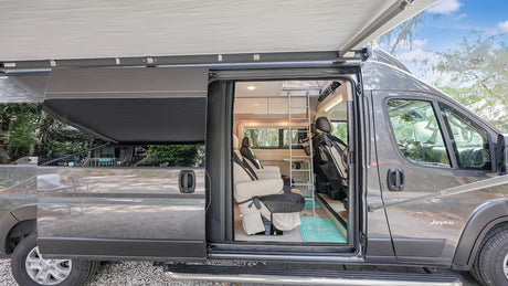 A gray RV rental with its side door open, showing the interior with swivel seats, ladder, and overhead awning.