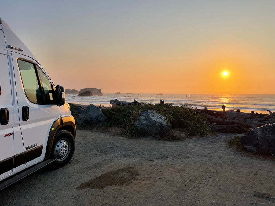 RV rental parked on a sandy beach access road, facing a vibrant orange sunset over the ocean with sea stacks.
