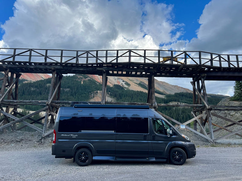  with colorful mountains & cloudy skies. Scenic travel.|Black RV rental parked by a scenic lake with mountains under a bright blue sky and fluffy clouds.|RV rental parked on gravel. A dark camper van with reflections overlooks red rock formations — photo 3