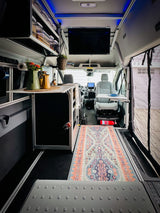 Explore a modern RV rental interior: functional kitchenette, swivel seat with table, smart overhead storage, and a colorful rug.