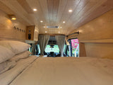 Cozy RV rental bedroom with warm wood paneling, comfy bed, and view to the front cabin, ready for adventure.