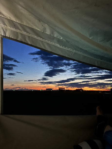 Relaxing inside an RV rental, admiring a stunning sunset through the window screen, showcasing a vibrant sky & distant horizon.