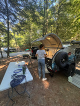 Woman using the outdoor kitchenette of an RV rental teardrop trailer at a wooded campsite. Other campers are seen nearby.