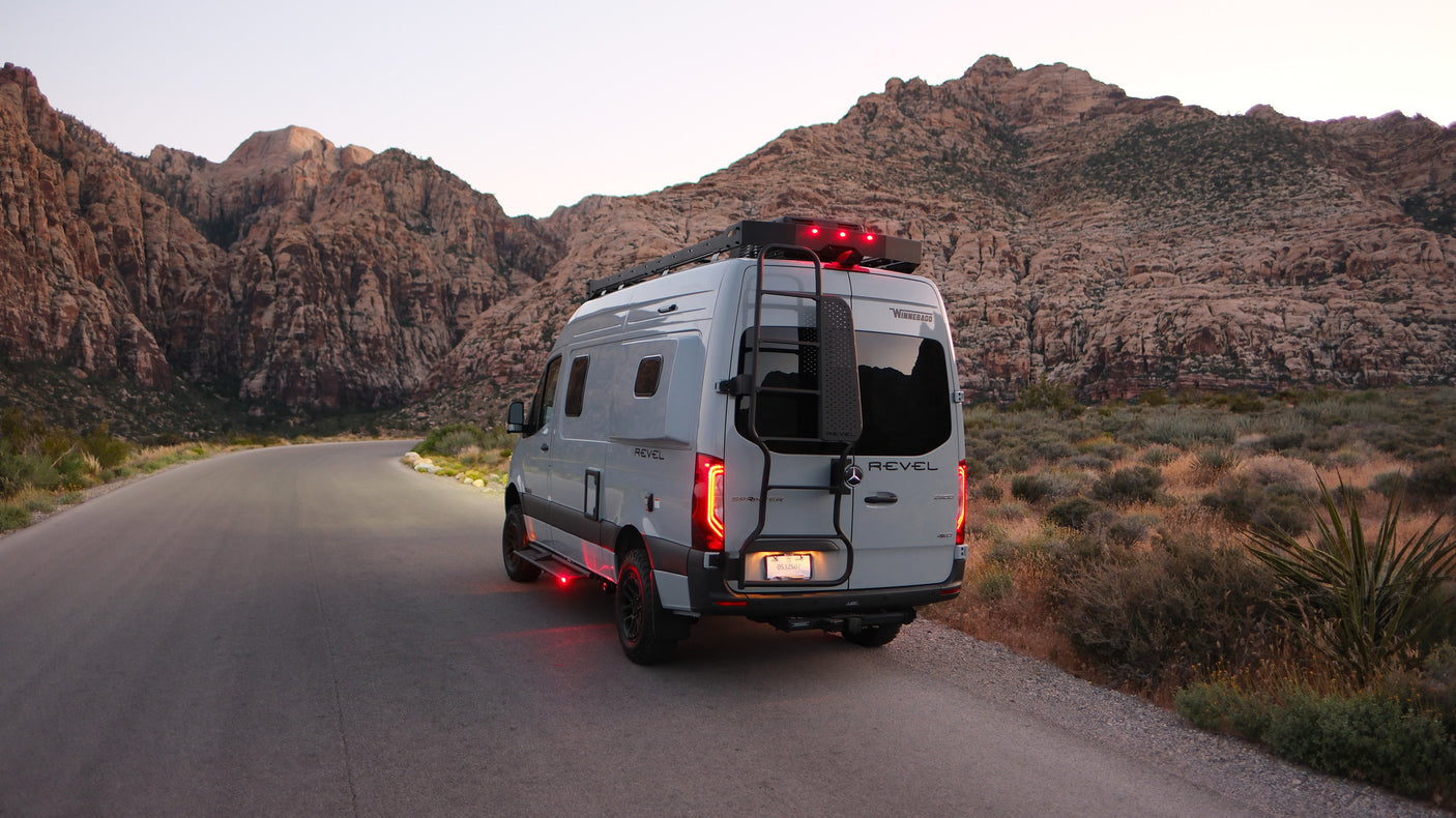 This grey Winnebago Revel RV rental explores scenic desert mountains at dusk, showcasing its red tail & underglow lights.