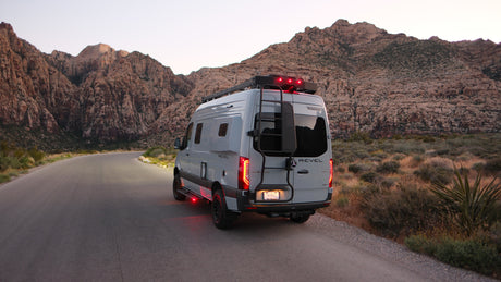 This grey Winnebago Revel RV rental explores scenic desert mountains at dusk, showcasing its red tail & underglow lights.