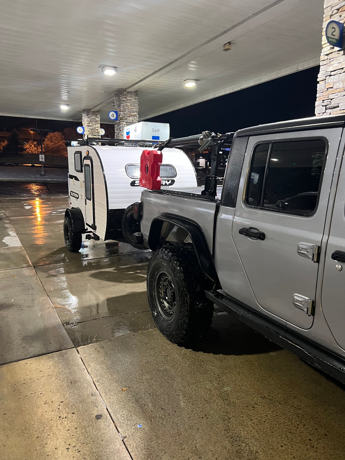 Off-road truck & teardrop RV rental at a gas station. Grey Jeep towing white camper, wet asphalt reflecting lights.