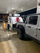Off-road truck & teardrop RV rental at a gas station. Grey Jeep towing white camper, wet asphalt reflecting lights.