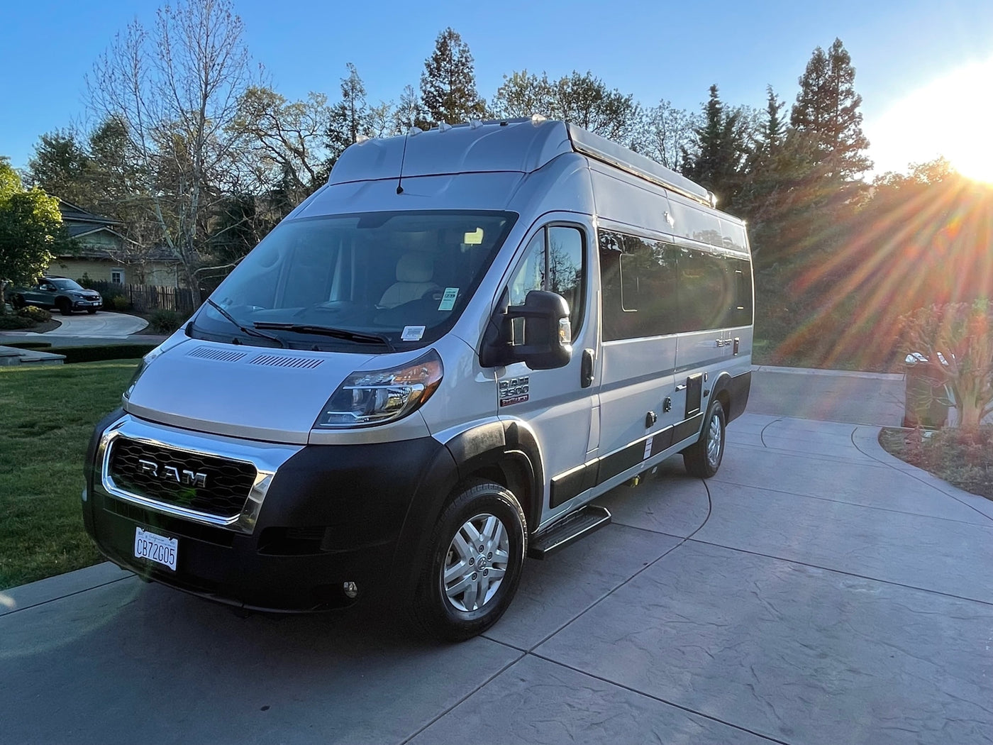 A silver Ram ProMaster rv rental, converted into a sleek camper van, sits on a driveway with sunrays. — photo 10