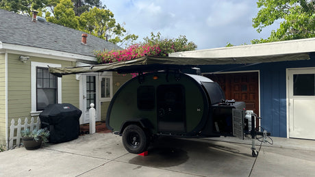 A dark green teardrop trailer with an extended awning is parked on a driveway between two houses. Ideal for rv rental camping.