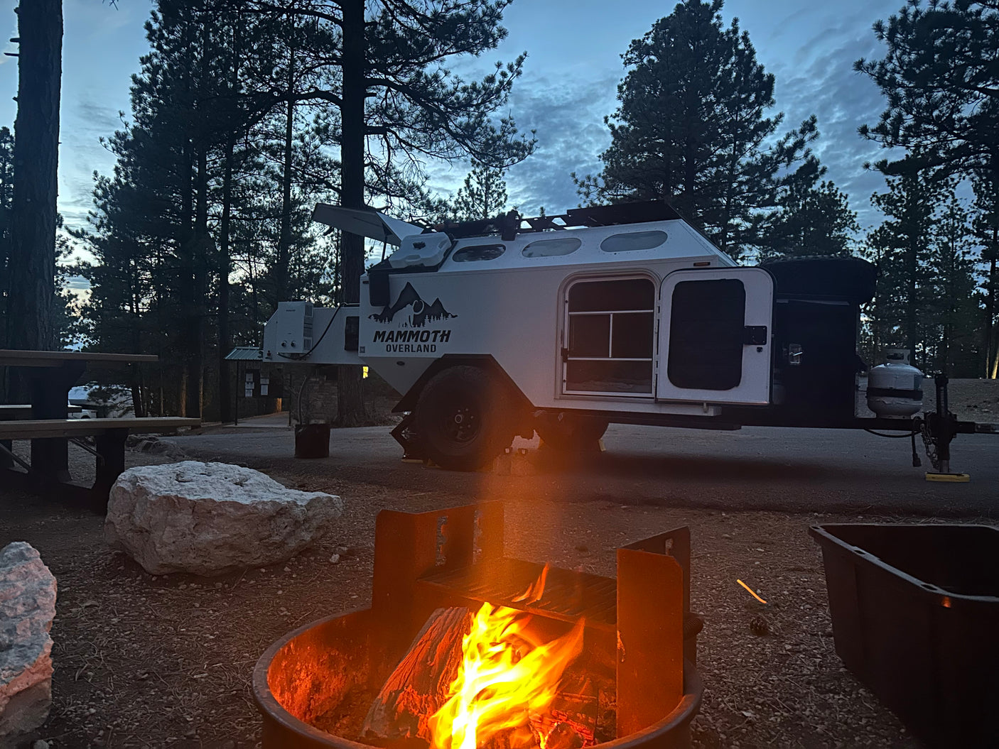 A cozy campfire burns brightly at a dusk campsite. A white overland RV rental trailer, doors open, is parked among pine trees.