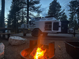 A cozy campfire burns brightly at a dusk campsite. A white overland RV rental trailer, doors open, is parked among pine trees.