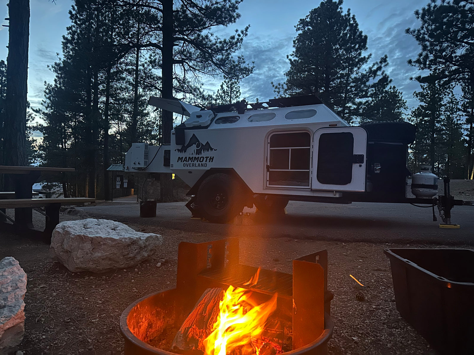 A cozy campfire burns brightly at a dusk campsite. A white overland RV rental trailer, doors open, is parked among pine trees.