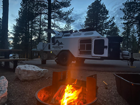 A cozy campfire burns brightly at a dusk campsite. A white overland RV rental trailer, doors open, is parked among pine trees.