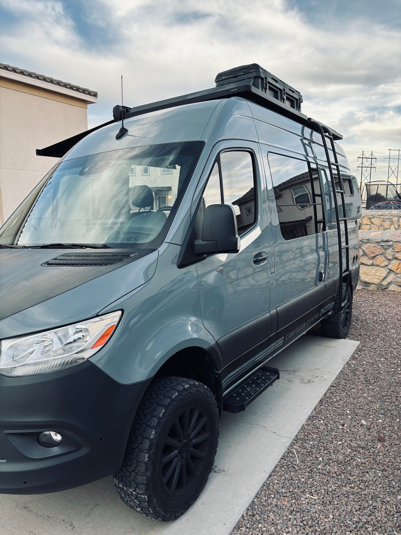 This grey off-road camper van, perfect for an RV rental, boasts a roof rack, side ladder, and rugged tires.