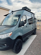 This grey off-road camper van, perfect for an RV rental, boasts a roof rack, side ladder, and rugged tires.