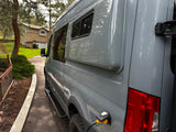 Grey RV rental van, seen from the side, charging via shore power. Parked on a driveway near a house and trees.