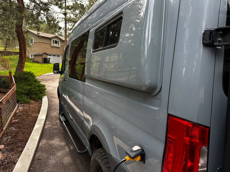 Grey RV rental van, seen from the side, charging via shore power. Parked on a driveway near a house and trees. — photo 9