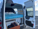 RV rental interior shows a comfy bed, storage, and open back doors revealing a serene water view.