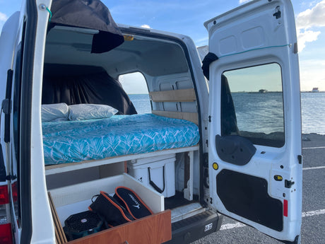 RV rental interior shows a comfy bed, storage, and open back doors revealing a serene water view.