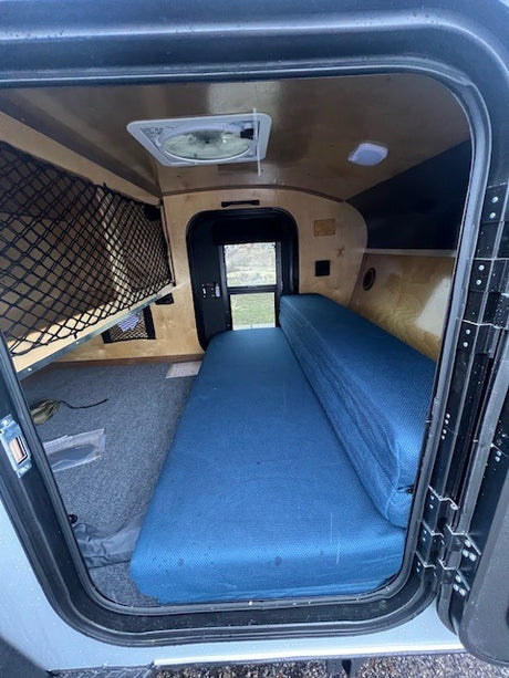 Compact RV rental interior with blue fold-out bed, mesh storage, ceiling fan, and back window.