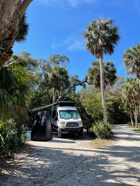A white Ford Transit camper van (rv rental) parked on a sandy path amidst lush palm trees in a sunny, tropical setting.