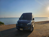 This dark Mercedes-Benz Sprinter **RV rental** sits on a sandy shore by serene blue water with sunlight reflecting.