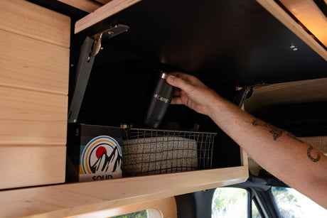 Hand retrieves travel mug from RV rental's overhead cabinet. Organized storage within a camper van.