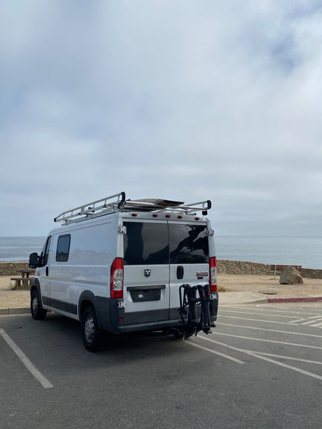 RV rental Ram ProMaster van with roof & bike racks by the ocean on a cloudy day, ready for coastal adventures.