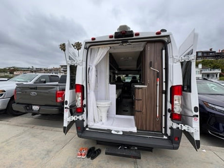 RV rental with open back doors, revealing a compact bathroom & cabinetry. Sandals rest on the ground on an overcast day.