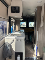 Interior of an rv rental, featuring a compact kitchen with a sink, overhead control panel, and an outdoor campsite view.