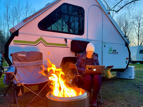 A woman sits bundled up, working on a laptop beside a bright campfire outside her A-liner RV rental at dusk.